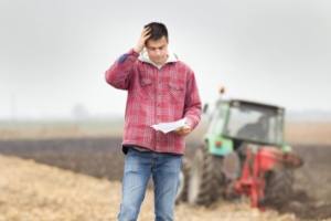 Man holding paperwork looking stressed, while someone operates a farm tiller in the background