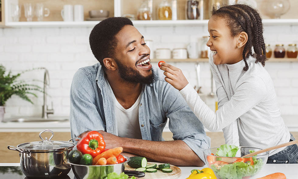 Daughter feeding father a tomato, while they make a salad